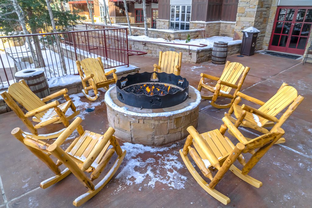 Wooden rocking chairs surround a lit fire pit in the winter.