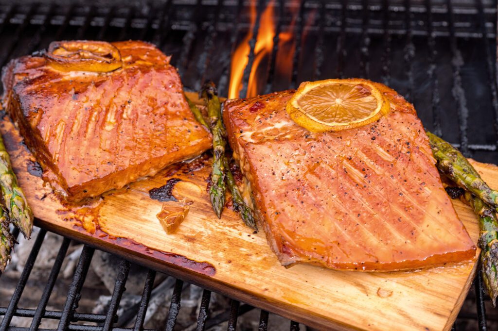 Pieces of salmon with lemons and asparagus cook on a wooden plank on a grill.