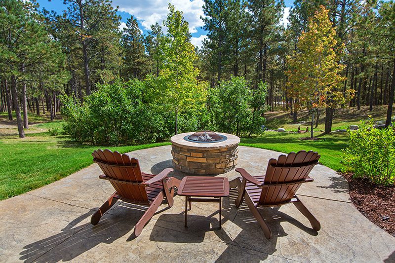Two wooden Adirondack chairs and a table are placed in front of a stone fire pit, overlooking a beautiful, wooded area.