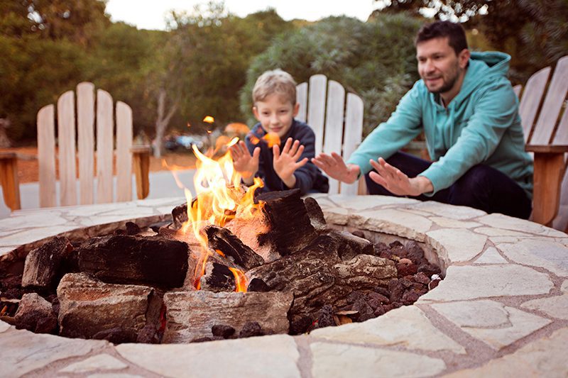 A father and young son warm their hands by a wood-burning fire pit.