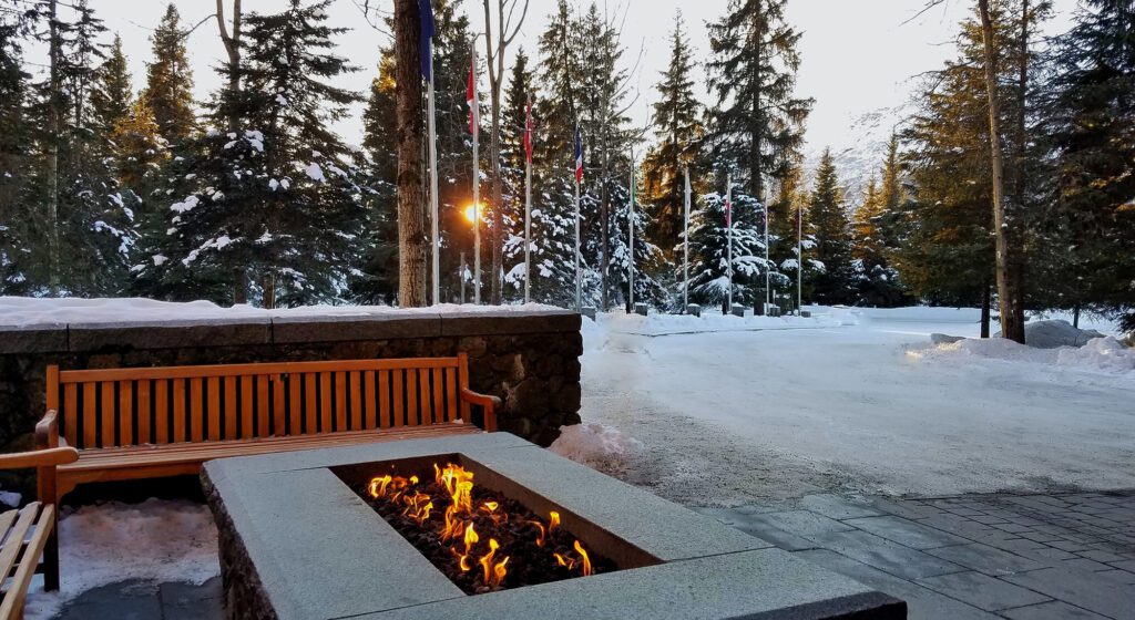 A fire table is lit on a stone patio with snow and trees in the background.