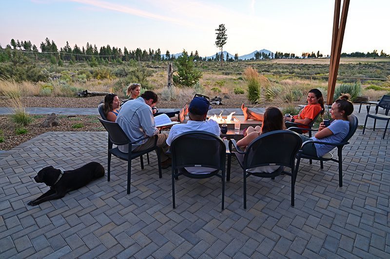 Friends gather around a fire pit table with a dog lounging on the side.