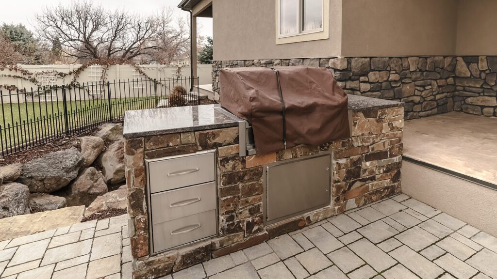 A grill cover and tarp strap is placed over a built-in grill on a natural stone outdoor kitchen.