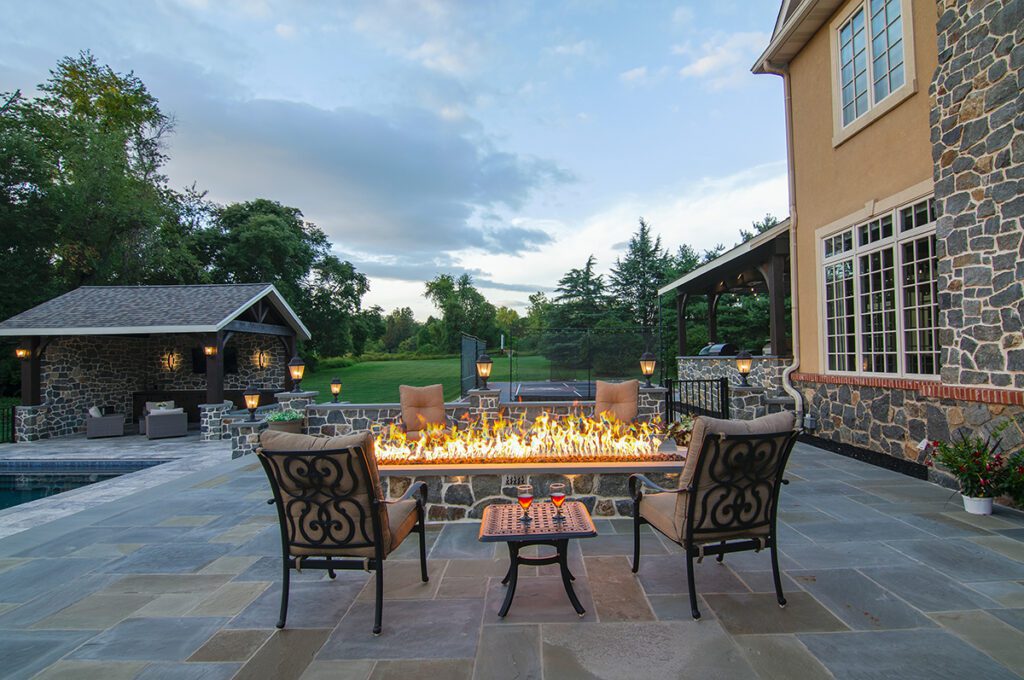 A long rectangle fire pit table burning with flames and surrounded by deck furniture with two glasses of wine on a side table.