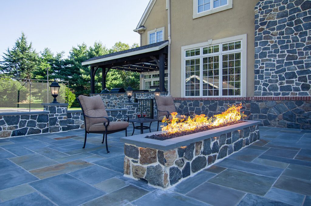 A long rectangle fire pit table burning with flames with two patio chairs next to it and two glasses of wine on a side table.