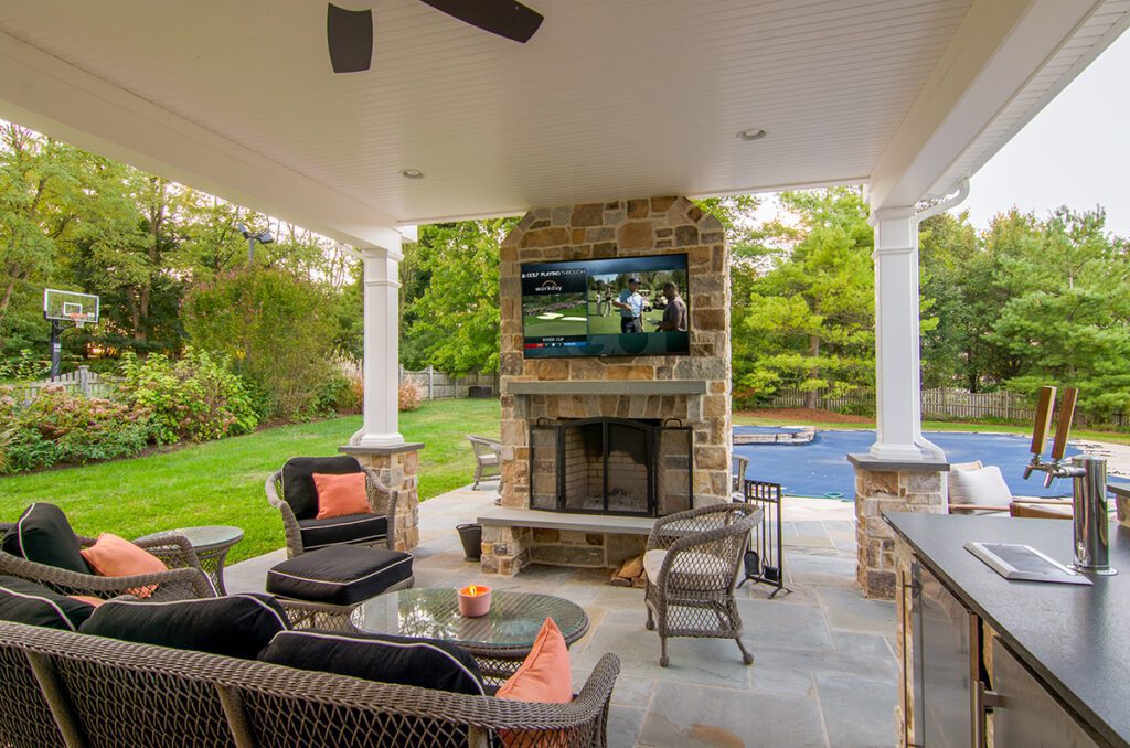 A backyard fireplace with a TV on the chimney surrounded by wicker furniture.