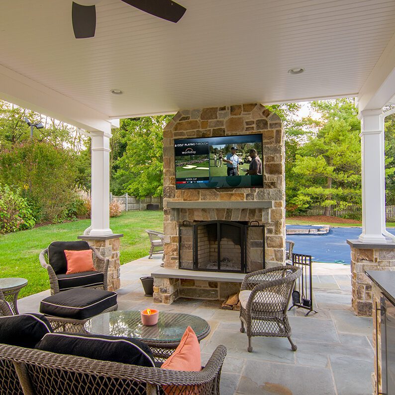An outdoor stone fireplace with a flat screen TV mounted and surrounded by wicker furniture with pillows.