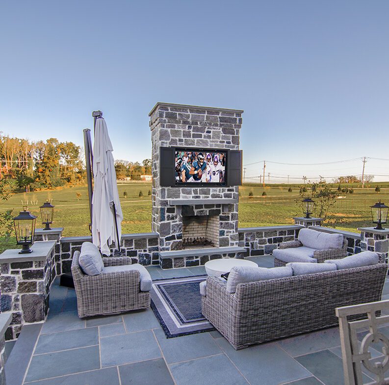 An outdoor stone patio with wicker furniture, a fireplace, and a flatscreen TV showing an Eagles football game.