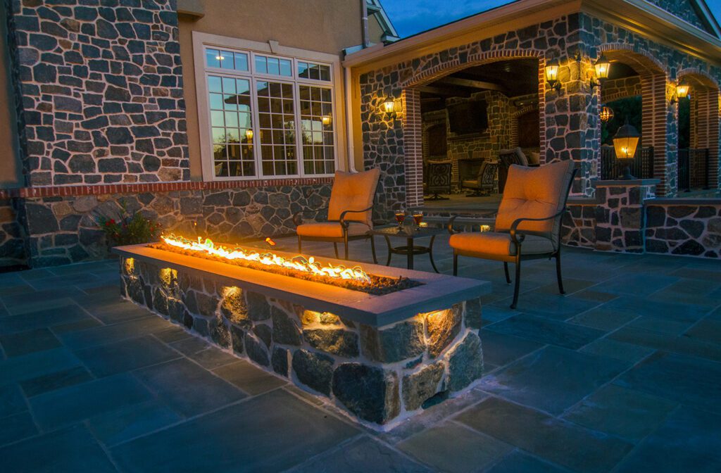 A long rectangular fire table with two chairs behind it on a stone patio.