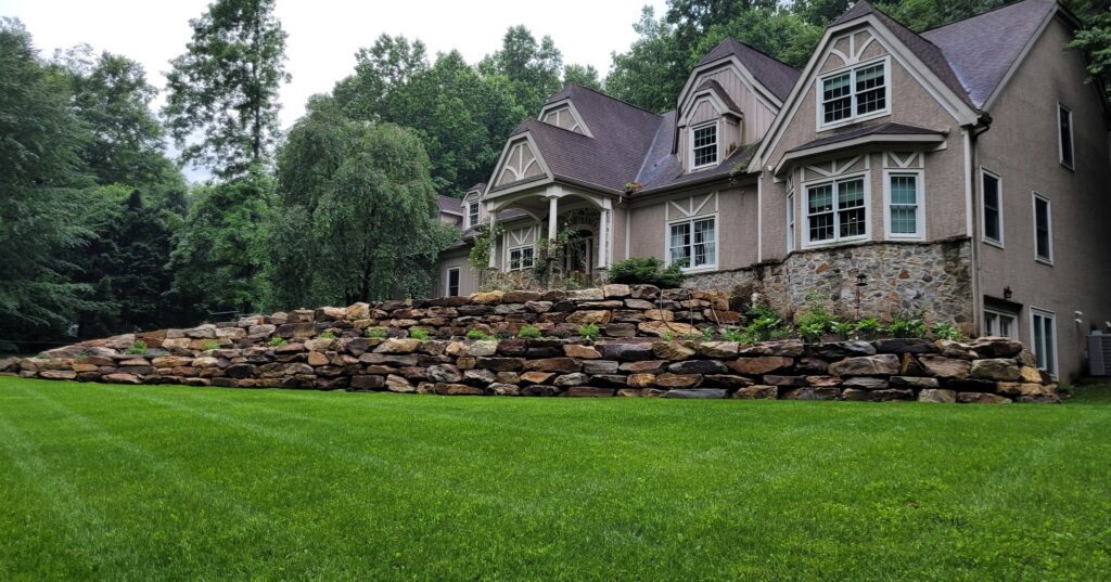 A stone retaining wall in between a green yard and a large home.